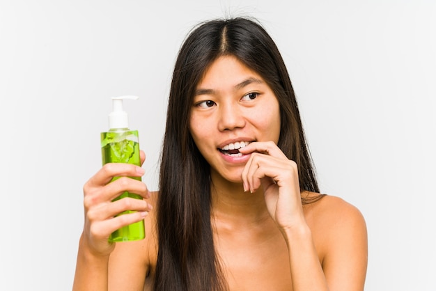 Jeune femme chinoise tenant une crème hydratante à l'aloe vera détendu en pensant à quelque chose en regardant un espace vide.