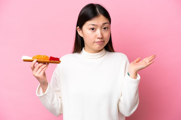 Jeune femme chinoise tenant sashimi isolé sur fond rose ayant des doutes tout en levant les mains