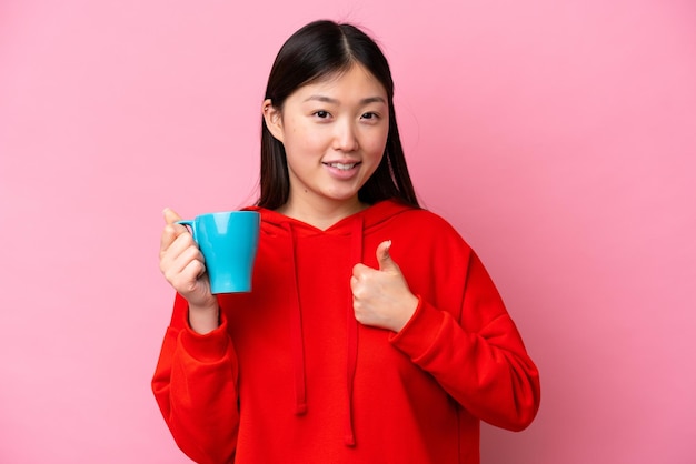 Jeune femme chinoise tenant une tasse de café isolée sur fond rose avec les pouces vers le haut parce que quelque chose de bien s'est produit