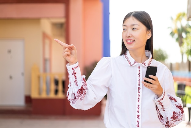 Jeune femme chinoise utilisant un téléphone portable à l'extérieur pointant vers le côté pour présenter un produit