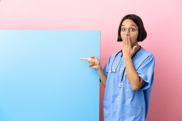 Jeune femme de chirurgien métisse avec une grande bannière sur fond isolé avec une expression de surprise tout en pointant le côté