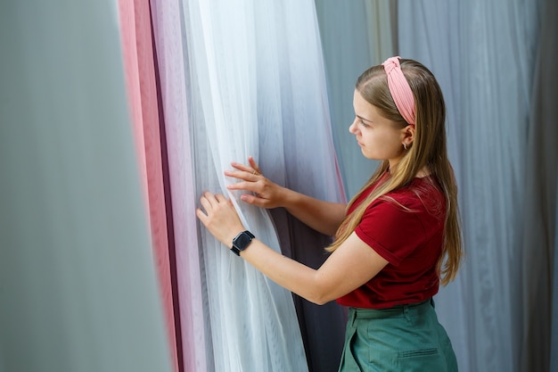 Jeune femme choisissant un tissu pour de nouveaux rideaux dans un magasin. Des échantillons du rideau sont suspendus à des cintres sur un rail dans le magasin. Échantillons de textures de tissu, de tulle et de rembourrage de meubles.