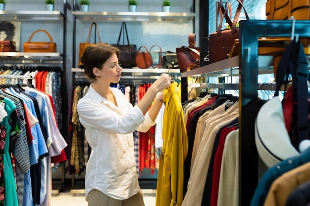 Jeune femme choisit l'imperméable dans un magasin de vêtements et tient un imperméable de couleur moutarde