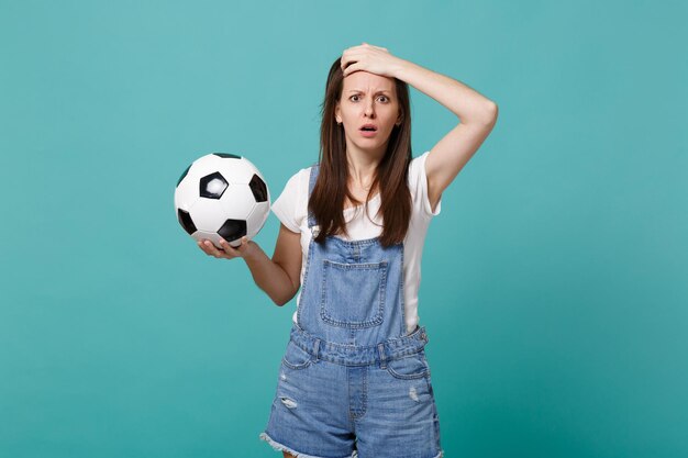 Une jeune femme choquée et abasourdie par un fan de football soutient l'équipe préférée avec un ballon de football mettant la main sur la tête isolée sur fond bleu turquoise. Émotions des gens, concept de mode de vie de loisirs familiaux sportifs.