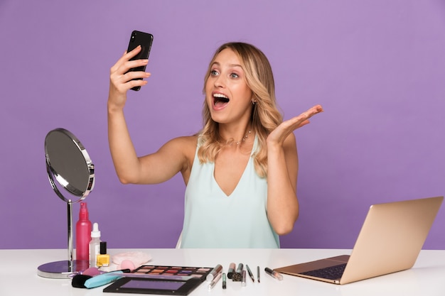 jeune femme choquée avec un ordinateur portable et des cosmétiques avec miroir parlant par téléphone portable.