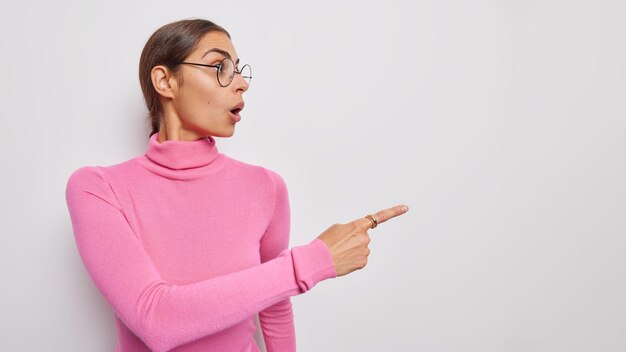 Une jeune femme choquée par ses émotions garde la bouche ouverte et dit que wow pointe le doigt juste à la promotion de la vente montre que la bannière de remise porte des lunettes rondes et un col roulé rose décontracté isolé sur un mur blanc