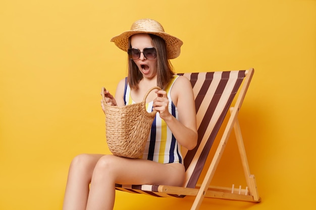 Une jeune femme choquée portant un maillot de bain rayé et un chapeau assis sur une chaise longue isolée sur fond jaune regardant à l'intérieur de son sac a oublié la crème solaire garde la bouche ouverte