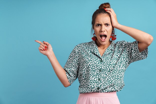 Jeune femme choquée portant des vêtements décontractés debout isolée sur un mur bleu, pointant du doigt l'espace de copie