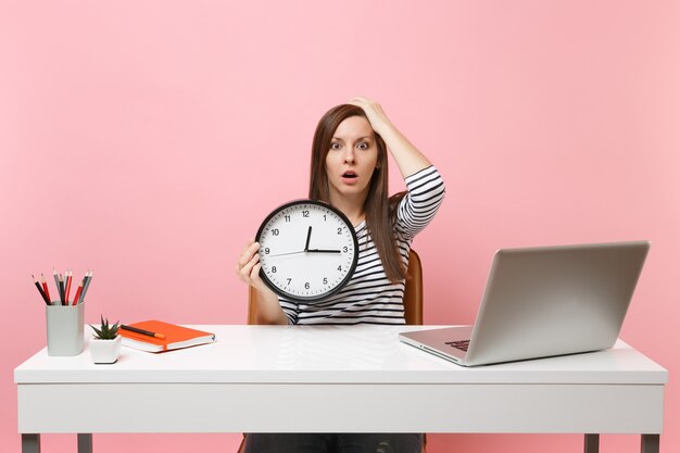 Une jeune femme choquée s'accrochant à la tête tient un réveil pendant qu'elle travaille au bureau avec un ordinateur portable