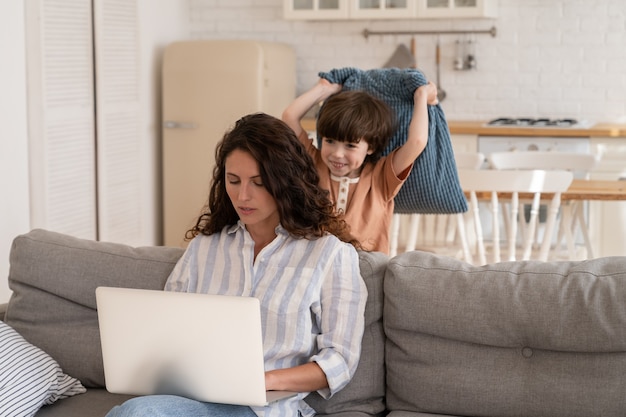 Une jeune femme ciblée utilise un ordinateur portable travaillant à domicile avec un fils d'enfant d'âge préscolaire l'a frappée avec un oreiller