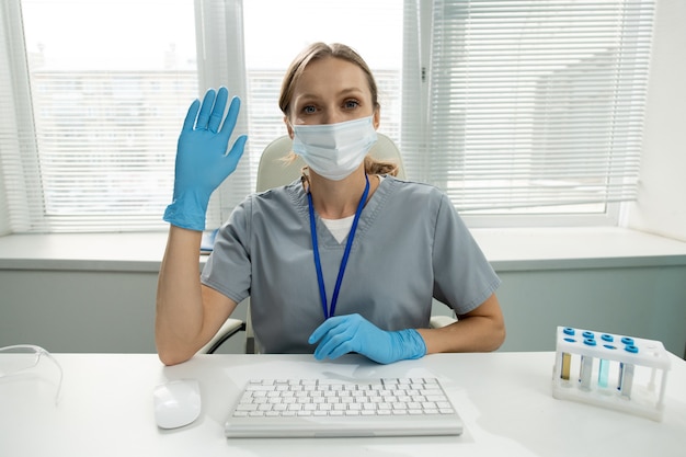 Jeune femme clinicienne ou travailleuse de laboratoire médical en vêtements de travail protecteurs vous montrant sa paume ouverte tout en gardant le coude sur la table