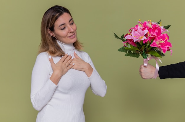 Jeune femme en col roulé blanc souriant main dans la main sur sa poitrine se sentant reconnaissante et positive des émotions tout en recevant le bouquet de fleurs