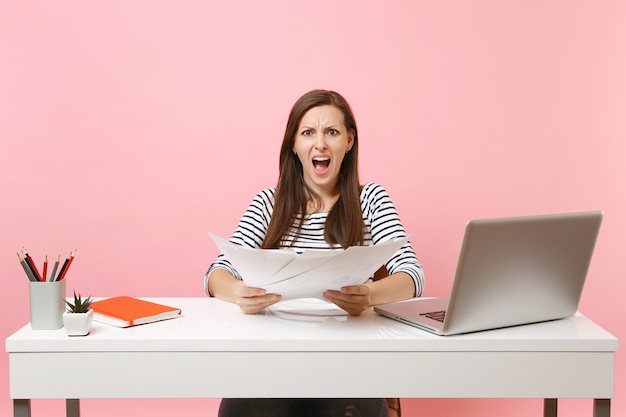 Jeune femme en colère ayant du mal à crier en tenant des documents papier, travaille sur un projet tout en étant assise au bureau avec un ordinateur portable