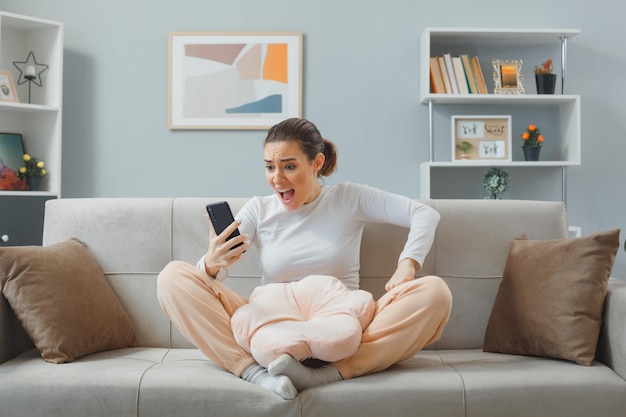 jeune femme en colère et frustrée en tenue décontractée assise sur un canapé à l'intérieur de la maison tenant un smartphone regardant l'écran sur son message de lecture mobile criant devenir fou