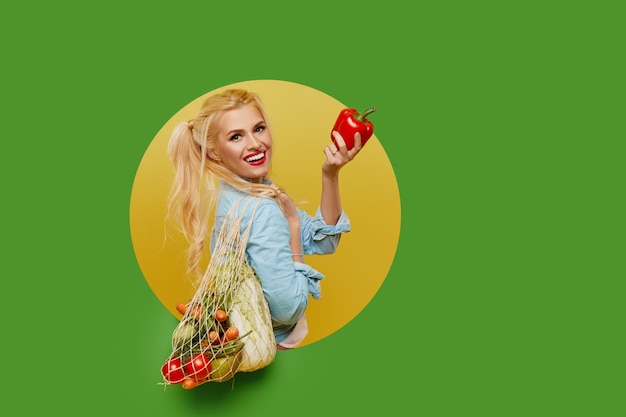 Jeune femme a collecté des légumes frais dans un sac de ficelle sur fond vert. Peeks hors d'un trou rond dans le mur.