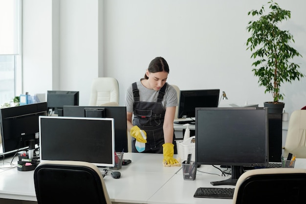 Jeune femme en combinaison et gants essuyant un bureau dans un grand bureau à espace ouvert