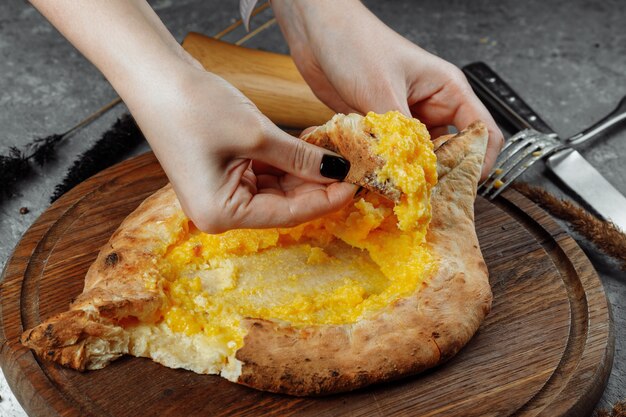 Une jeune femme commence à manger du khachapuri parfumé en ajarian avec du fromage sur un support en bois avec un