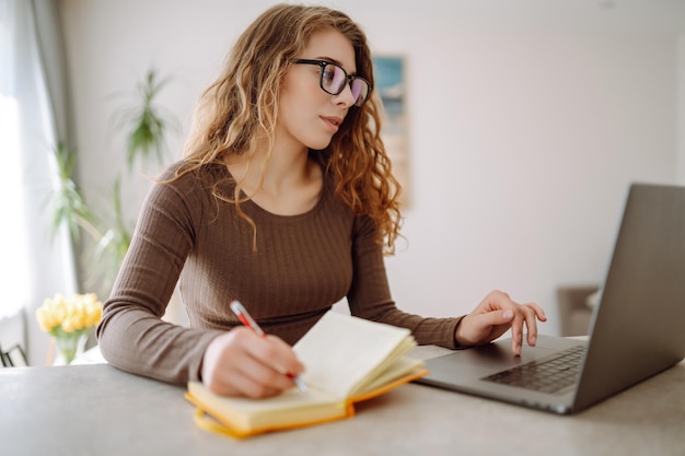 Une jeune femme concentrée est assise à table avec un ordinateur portable et un bloc-notes lumineux.