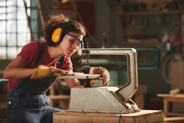 Jeune femme concentrée en lunettes de protection et cache-oreilles faisant des trous dans une planche en bois avec une perceuse à colonne