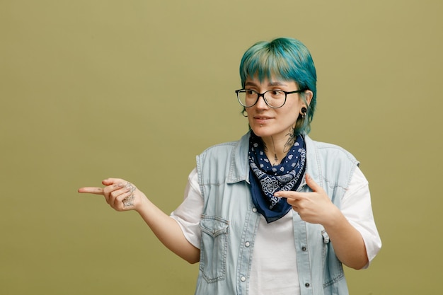 Jeune femme concentrée portant des lunettes bandana sur le cou regardant le côté pointant vers le côté isolé sur fond vert olive