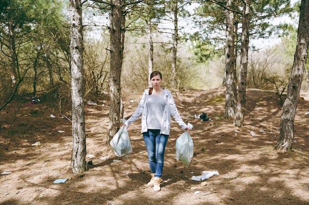 Jeune femme concernée choquée dans des vêtements décontractés tenant des sacs poubelle nettoyant les ordures dans un parc ou une forêt jonché de déchets. Problème de pollution de l'environnement. Arrêtez les ordures de la nature, concept de protection de l'environnement.