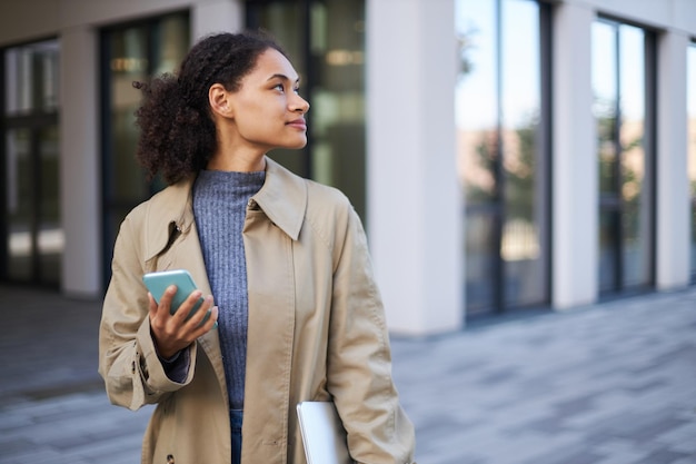 Jeune femme confiante afro-américaine avec téléphone portable et ordinateur portable contre un immeuble de bureaux moderne