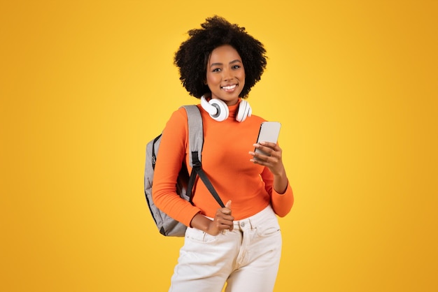 Une jeune femme confiante avec des cheveux afro bouclés et des écouteurs autour du cou vérifie son smartphone