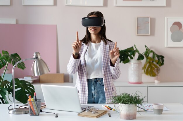 Jeune femme confiante dans un casque de réalité virtuelle pointant en l'air en se tenant debout sur son lieu de travail au bureau