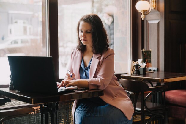 Une jeune femme confiante dans un costume d'affaires travaille sur un ordinateur portable tout en étant assise près de la fenêtre dans un café