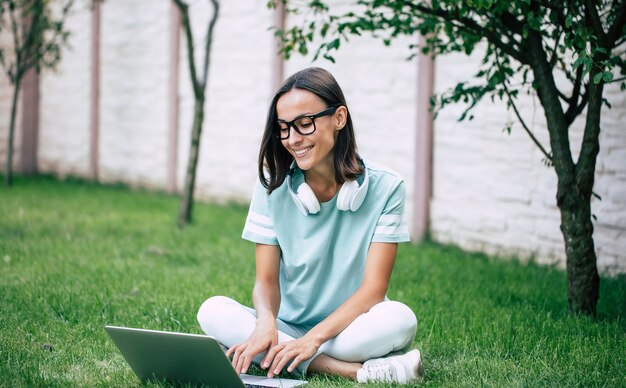 Jeune femme confiante heureuse belle moderne dans des écouteurs et des lunettes travaille sur un ordinateur portable alors qu'il était assis sur la pelouse.