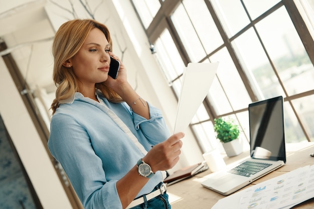 Jeune femme confiante parlant au téléphone et regardant un document tout en travaillant au bureau