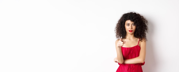 Jeune femme confiante en robe rouge et maquillage pointant vers l'appareil photo en vous choisissant d'inviter à l'événement stan