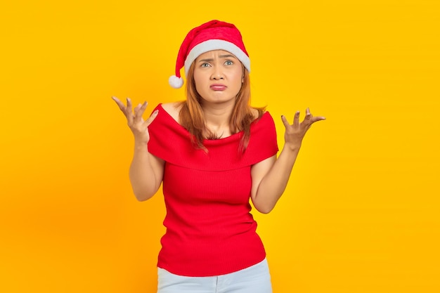 Jeune femme confuse portant un chapeau de Noël haussant les épaules et les mains sur fond jaune