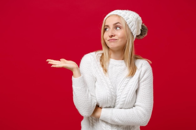 Jeune femme confuse en pull blanc chapeau isolé sur fond rouge portrait en studio. Concept de saison froide d'émotions de personnes de mode de vie sain. Maquette de l'espace de copie. Regardant de côté en écartant les mains.