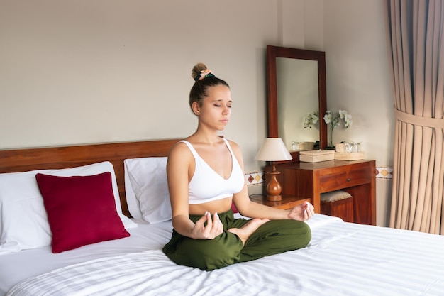 Jeune femme consciente médite tenant les doigts dans le signe du yoga assis sur un lit blanc dans la chambre