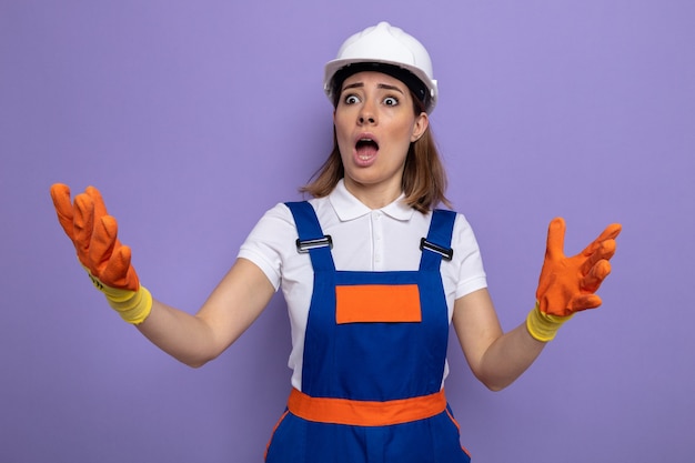 Jeune femme de construction en uniforme de construction et casque de sécurité dans des gants en caoutchouc regardant de côté, confus et inquiet, écartant les bras sur les côtés, debout sur un mur violet