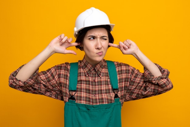 Jeune femme de construction en uniforme de construction et casque de sécurité fermant les oreilles avec les doigts avec une expression agacée debout sur orange