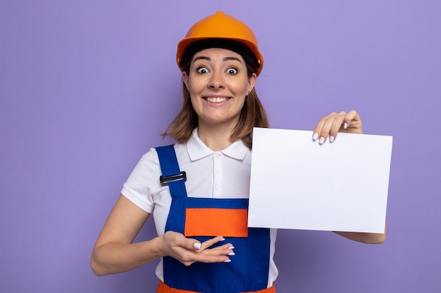 Jeune femme de construction en uniforme de construction et casque de sécurité tenant une page blanche présentant le bras souriant joyeusement debout sur violet