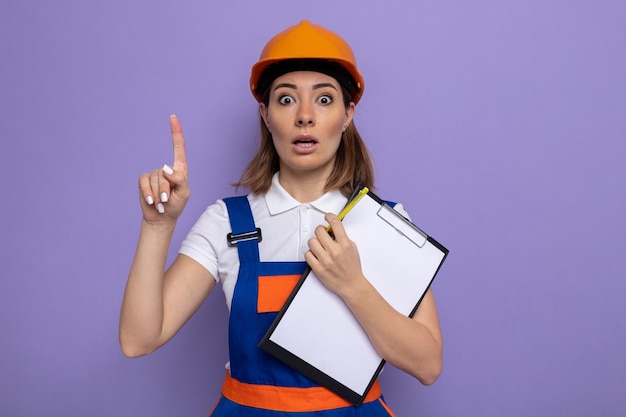 Jeune femme de construction en uniforme de construction et casque de sécurité tenant un presse-papiers avec des pages blanches l'air surpris montrant l'index ayant une nouvelle idée debout sur un mur violet