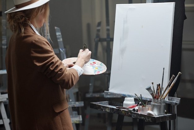 Jeune femme contemporaine avec palette de couleurs mélangeant les couleurs tout en allant peindre sur chevalet avec du papier blanc en studio