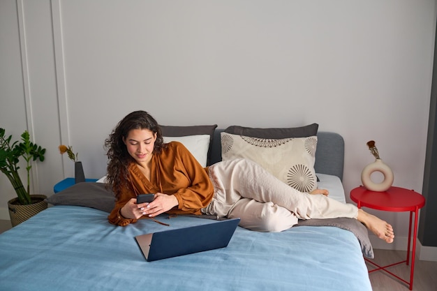 Jeune femme contemporaine avec smartphone reposant sur un lit double