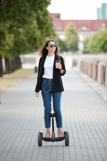 Jeune femme contemporaine avec un verre de café regardant la rivière tout en roulant sur un gyroscope le long de la route contre des arbres et des bâtiments verts