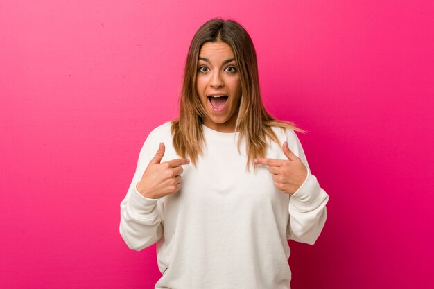 Jeune femme contre un mur surpris pointant du doigt, souriant largement.