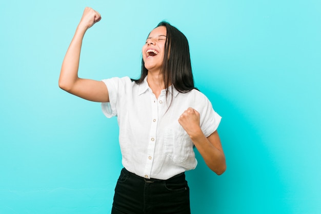 Jeune femme cool hispanique contre un mur bleu, levant le poing après une victoire, concept gagnant.