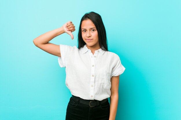 Jeune Femme Cool Hispanique Contre Un Mur Bleu Montrant Un Geste D'aversion, Pouce En Bas. Concept De Désaccord.