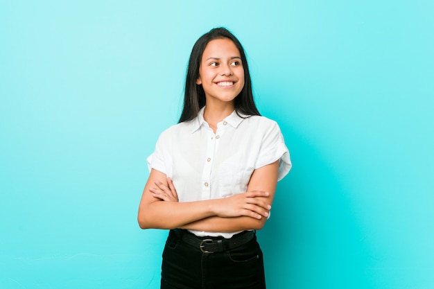 Jeune femme cool hispanique contre un mur bleu souriant confiant avec les bras croisés.