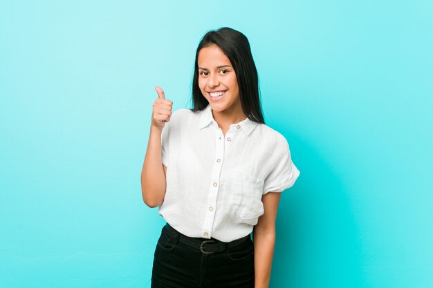 Jeune femme cool hispanique contre un mur bleu souriant et levant le pouce vers le haut