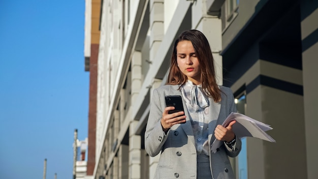 Jeune femme en costume d'affaires avec un téléphone et des documents marchant dans la rue.