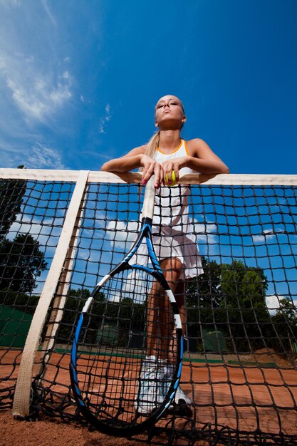 Jeune femme en costume blanc et raquette de tennis dans ses mains debout derrière le filet. Vue de dessous