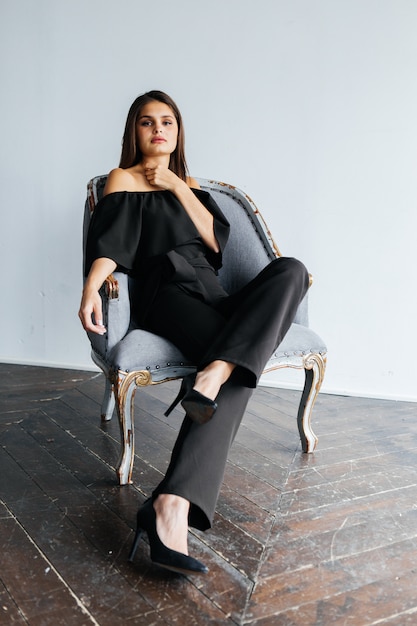 Jeune femme en costume noir assis sur une chaise à l'intérieur clair. Femme d'affaires. Belle femme